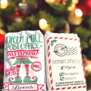 May include: Two white Christmas gift bags with red and green trim. The bag on the left has a green elf illustration and the text "North Pole Post Office Elf Express" and "Lucas". The bag on the right has the text "North Pole Express Delivery" and "Overnight Service For Elijah Santa Approved Checked 2x If Undeliverable, Return To: Santa Claus, The North Pole".