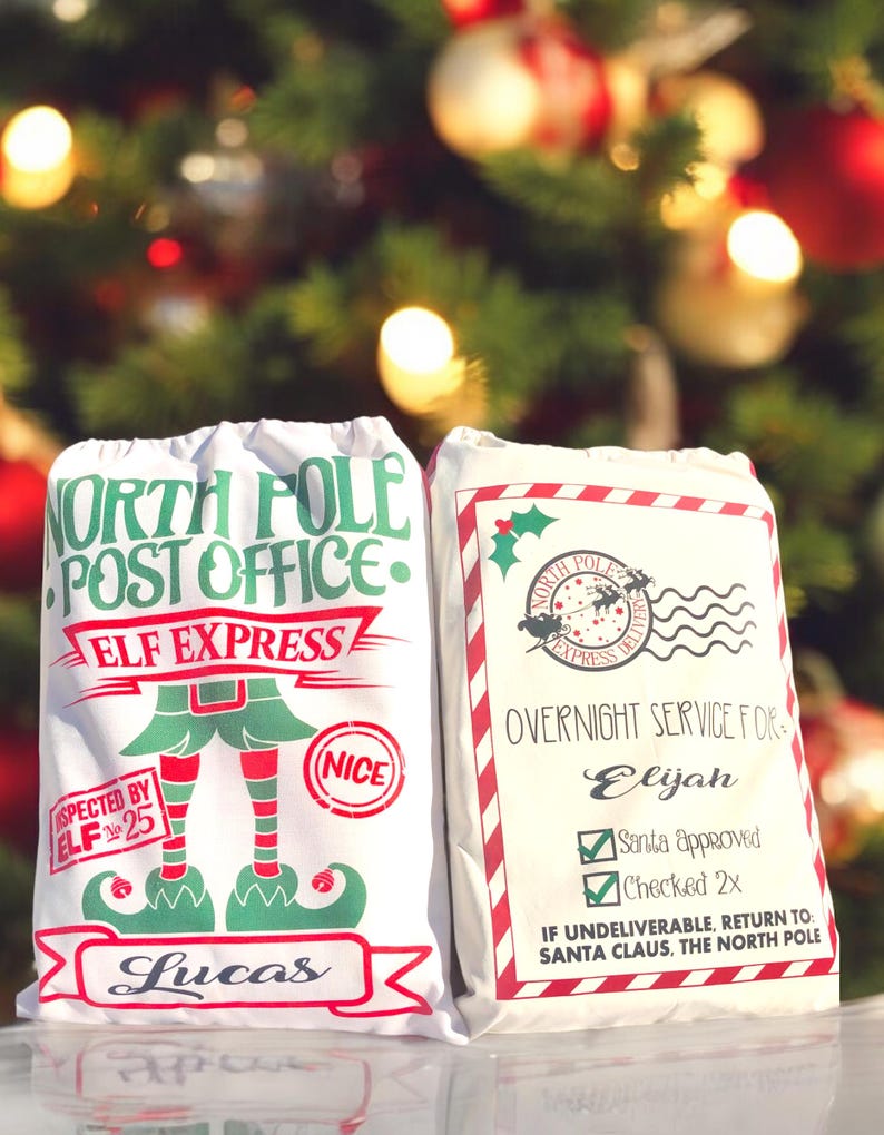 May include: Two white Christmas gift bags with red and green trim. The bag on the left has a green elf illustration and the text "North Pole Post Office Elf Express" and "Lucas". The bag on the right has the text "North Pole Express Delivery" and "Overnight Service For Elijah Santa Approved Checked 2x If Undeliverable, Return To: Santa Claus, The North Pole".