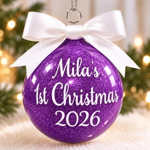 May include: A spherical purple glitter Christmas ornament featuring a white satin ribbon bow at the top. The ornament is printed with the white text "Mila's 1st Christmas 2026" in a script and sans-serif font combination, suspended by a silver metal loop.