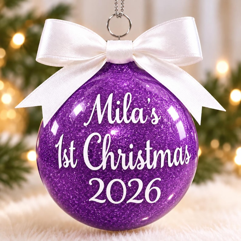 May include: A spherical purple glitter Christmas ornament featuring a white satin ribbon bow at the top. The ornament is printed with the white text "Mila's 1st Christmas 2026" in a script and sans-serif font combination, suspended by a silver metal loop.