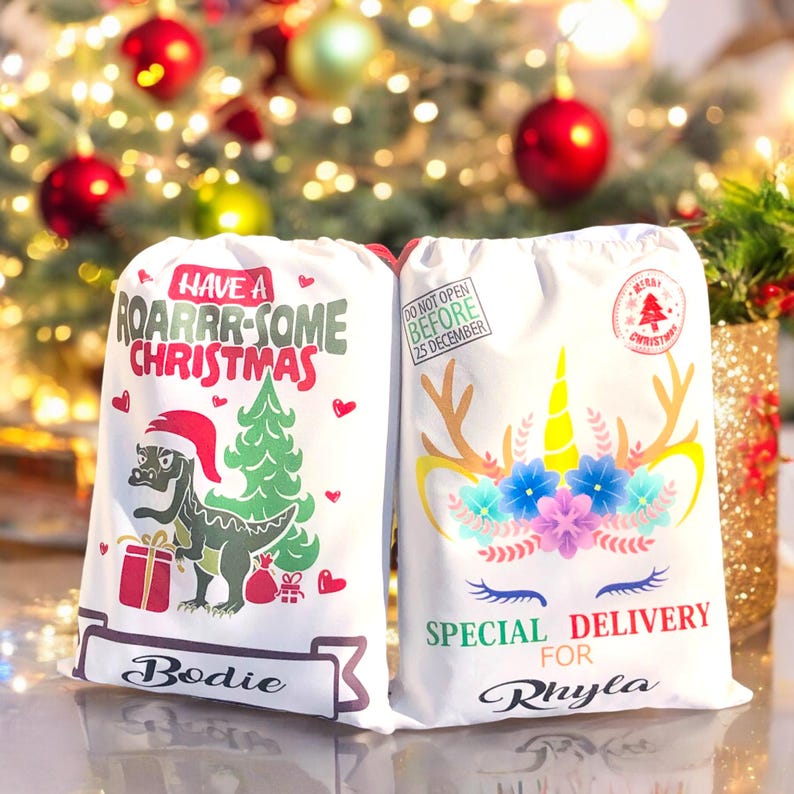 May include: Two white Christmas gift bags with personalized names. One bag features a dinosaur with the text "Have a Roarrr-some Christmas" and the other features a unicorn with the text "Special Delivery for".