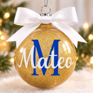 May include: A clear spherical Christmas ornament filled with gold glitter, featuring a large blue letter M and the name Mateo in white script. The ornament is topped with a silver cap and a tied white satin ribbon bow for hanging.