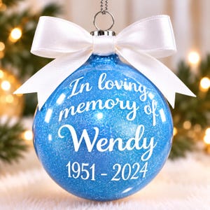 May include: A round blue glitter Christmas ornament featuring white text that reads In loving memory of Wendy 1951-2024. The ornament is topped with a silver metal cap and a white satin ribbon tied in a bow, hanging from a silver string.