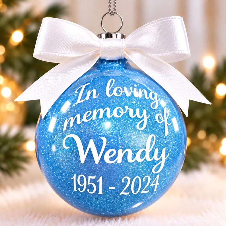 May include: A round blue glitter Christmas ornament featuring white text that reads In loving memory of Wendy 1951-2024. The ornament is topped with a silver metal cap and a white satin ribbon tied in a bow, hanging from a silver string.