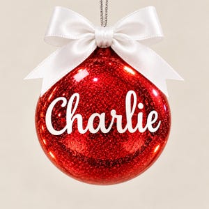 May include: A spherical red Christmas ornament with a glittery finish, featuring the name Charlie written in white script lettering across the center. The ornament is topped with a smooth white satin ribbon tied into a bow and a silver hanging cord.