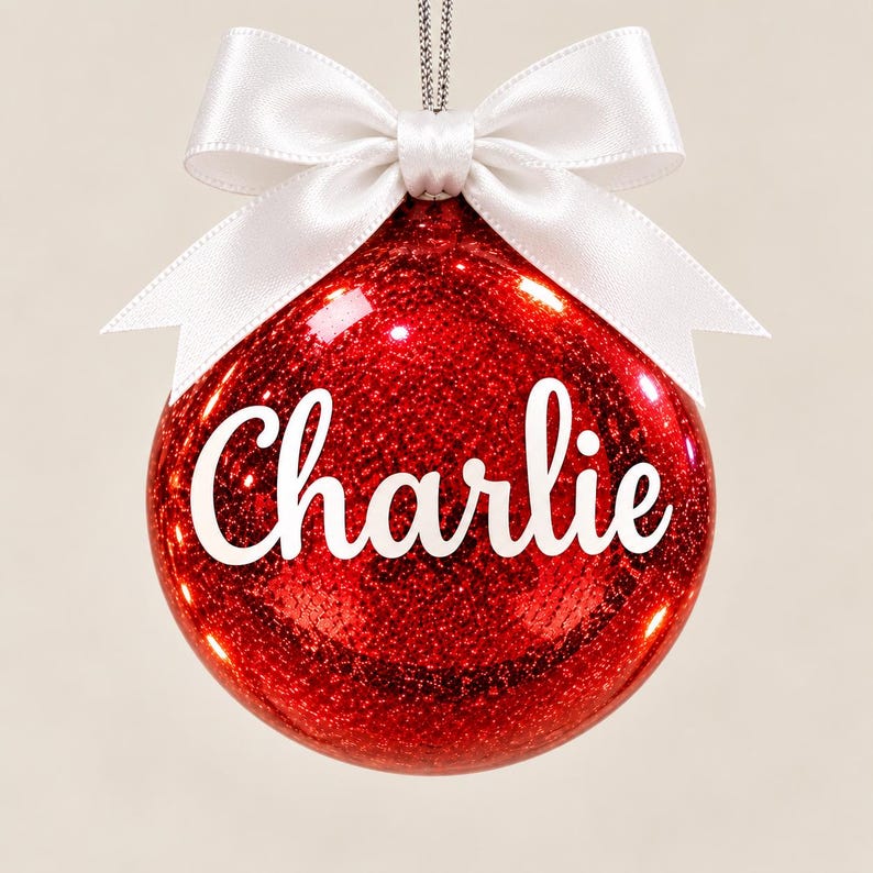 May include: A spherical red Christmas ornament with a glittery finish, featuring the name Charlie written in white script lettering across the center. The ornament is topped with a smooth white satin ribbon tied into a bow and a silver hanging cord.