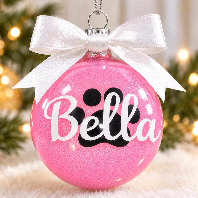 May include: A spherical pink glitter Christmas ornament featuring a black paw print graphic and the name Bella written in white script lettering. The ornament is topped with a silver metal cap and a decorative white satin ribbon bow.