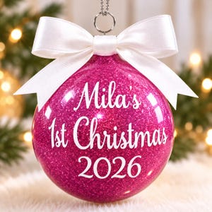 May include: A spherical, hot pink glitter Christmas ornament featuring the white text "Mila's 1st Christmas 2026" printed on the front. The ornament is topped with a silver metal hanging loop and a large, crisp white satin ribbon bow.