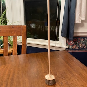 May include: A wooden dowel rod with a natural wood finish is inserted into a wooden base. The base is a round slice of wood with a natural bark texture.
