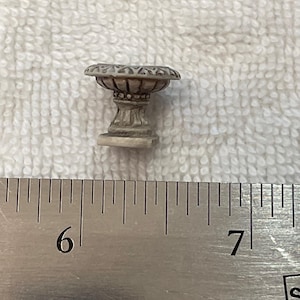 May include: A miniature, off-white, decorative planter with a detailed, ornate design. The planter has a round bowl shape with a patterned rim, supported by a pedestal base. The planter is approximately 1 inch tall.