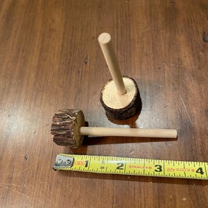 May include: Two wooden dowels with a round wooden base. The dowels are approximately 3 inches long. A yellow measuring tape is in the foreground.