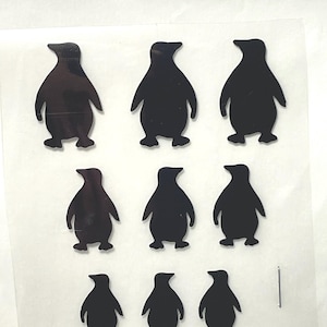 May include: Nine black penguin silhouettes on a clear plastic sheet. The penguins are of varying sizes and are arranged in three rows.