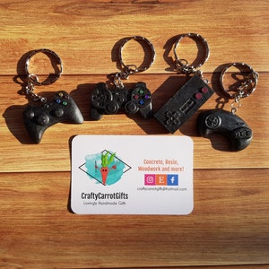 Puede incluir: Cuatro llaveros de resina negra en forma de mandos de juego con anillas de plata. Los llaveros tienen forma de diferentes mandos de juego, incluyendo un mando de Playstation, un mando de Xbox, un mando de Nintendo y un mando genérico. Los llaveros están sobre una superficie de madera con una tarjeta de visita que dice "CraftyCarrotGifts Lovingly Handmade Gifts Concrete, Resin, Woodwork and more!"