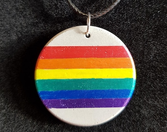 Puede incluir: Un collar redondo de color blanco con un diseño de arcoíris pintado. El colgante está hecho de hormigón y tiene un cordón negro.