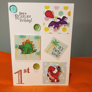 Op de afbeelding: Een witte kaart met een dinosaurus thema voor een eerste verjaardag. De kaart heeft een groene dinosaurus, een paarse dinosaurus, een rode dinosaurus en de tekst "Have a ROARING Birthday!" en "It's party time!"