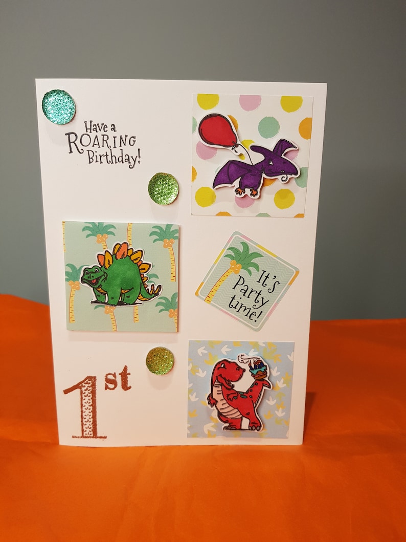 Op de afbeelding: Een witte kaart met een dinosaurus thema voor een eerste verjaardag. De kaart heeft een groene dinosaurus, een paarse dinosaurus, een rode dinosaurus en de tekst "Have a ROARING Birthday!" en "It's party time!"