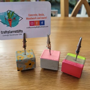 Puede incluir: Tres porta tarjetas de hormigón y pintados con clips metálicos. Los soportes están pintados en diferentes colores y patrones, incluyendo amarillo, rosa, verde y azul. Los porta tarjetas están sobre una superficie de madera. El porta tarjetas de la izquierda tiene un patrón floral, el porta tarjetas del medio es rosa y el porta tarjetas de la derecha tiene rayas verdes y rosas.