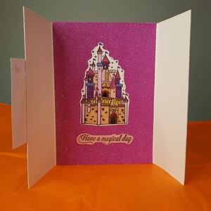 Op de afbeelding: Een roze glitterkaart met een cartoon-kasteel illustratie en de tekst "Have a magical day".