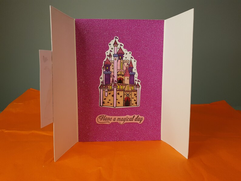 Op de afbeelding: Een roze glitterkaart met een cartoon-kasteel illustratie en de tekst "Have a magical day".