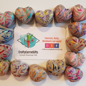 May include: A collection of 15 small, gray concrete heart-shaped ornaments. Each heart is decorated with colorful, abstract patterns in shades of pink, blue, green, gold, and orange. The hearts are arranged in a circle around a business card with the text "CraftyCarrotGifts" and "Lovingly Handmade Gifts".