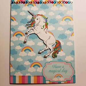 Op de afbeelding: Een kaart met een regenboog-eenhoorn op een blauwe achtergrond met wolken en regenbogen. De kaart heeft een roze rand en een wit banner met de tekst "Have a magical day".