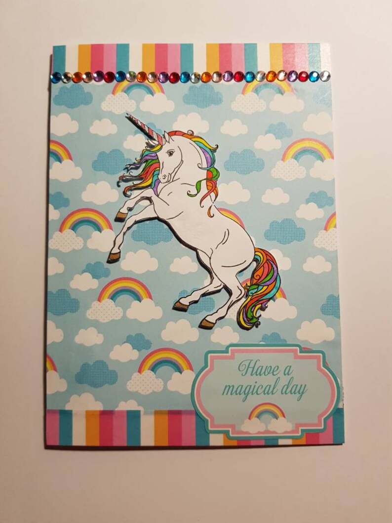 Op de afbeelding: Een kaart met een regenboog-eenhoorn op een blauwe achtergrond met wolken en regenbogen. De kaart heeft een roze rand en een wit banner met de tekst "Have a magical day".