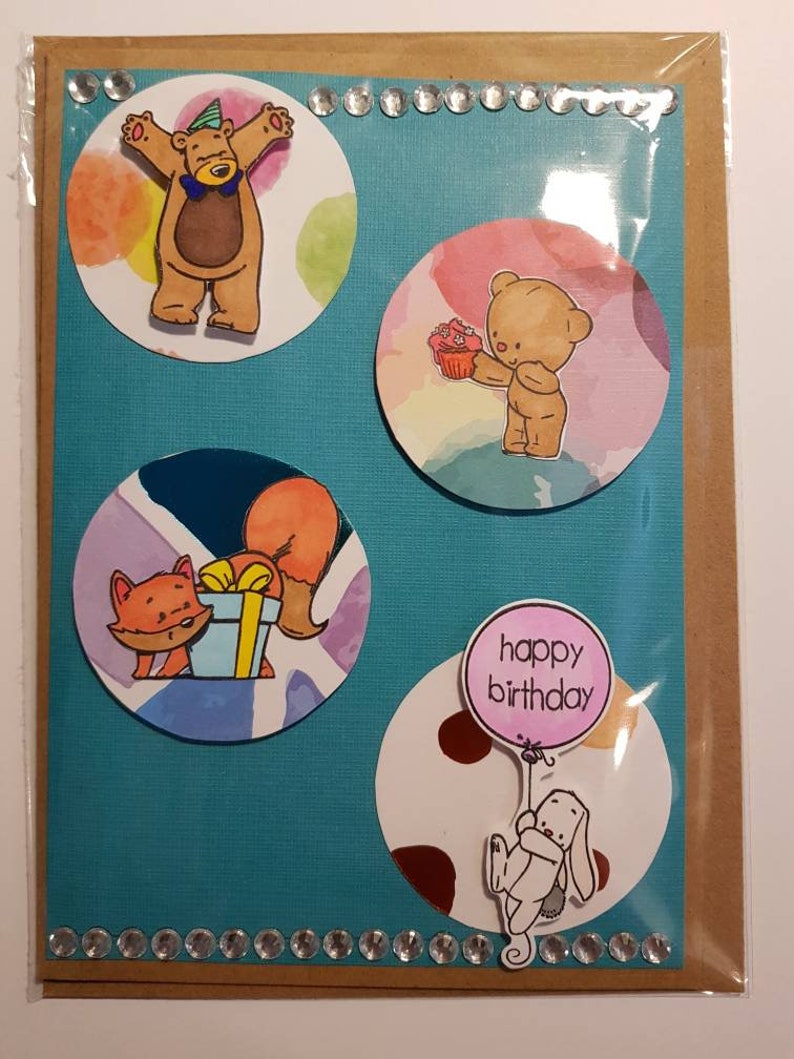 Op de afbeelding: Een handgemaakte verjaardagskaart met een blauwe achtergrond en vier ronde uitsparingen. De uitsparingen tonen illustraties van een beer met een feestmuts, een beer die een cupcake vasthoudt, een vos die een cadeau vasthoudt en een konijn dat een ballon vasthoudt waarop "happy birthday" staat.