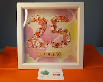 May include: A framed artwork featuring the number 25 made from colorful buttons, with the name "KARLA" spelled out in Scrabble tiles below. The background is a watercolor-style design in shades of pink and yellow.