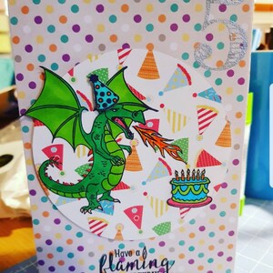Op de afbeelding: Een verjaardagskaart met een groene draak die vuur spuit, een blauwe en witte taart met kaarsen en de tekst "Have a flaming good birthday". De kaart heeft een stippenachtergrond met een zilveren glittercijfer 5.