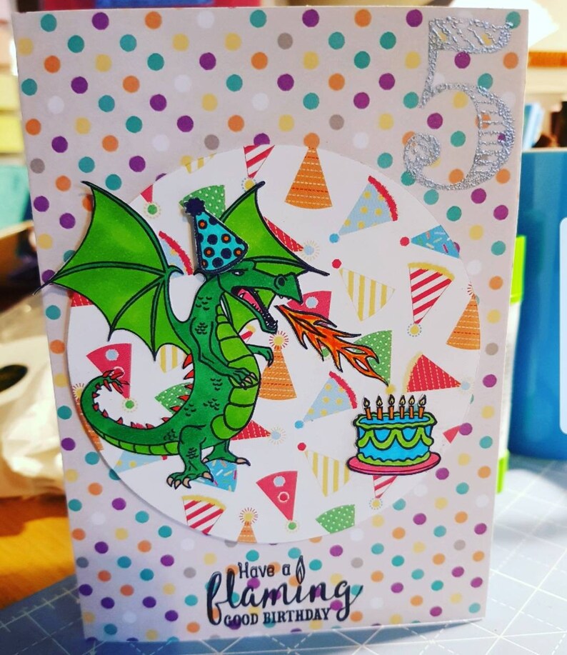 Op de afbeelding: Een verjaardagskaart met een groene draak die vuur spuit, een blauwe en witte taart met kaarsen en de tekst "Have a flaming good birthday". De kaart heeft een stippenachtergrond met een zilveren glittercijfer 5.