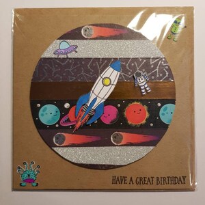 Op de afbeelding: Een bruine kaart met een cirkel in het midden. De cirkel is gevuld met een ruimte sc&egrave;ne met een raket, planeten, sterren en een UFO. De tekst "HAVE A GREAT BIRTHDAY" staat onderaan de kaart.