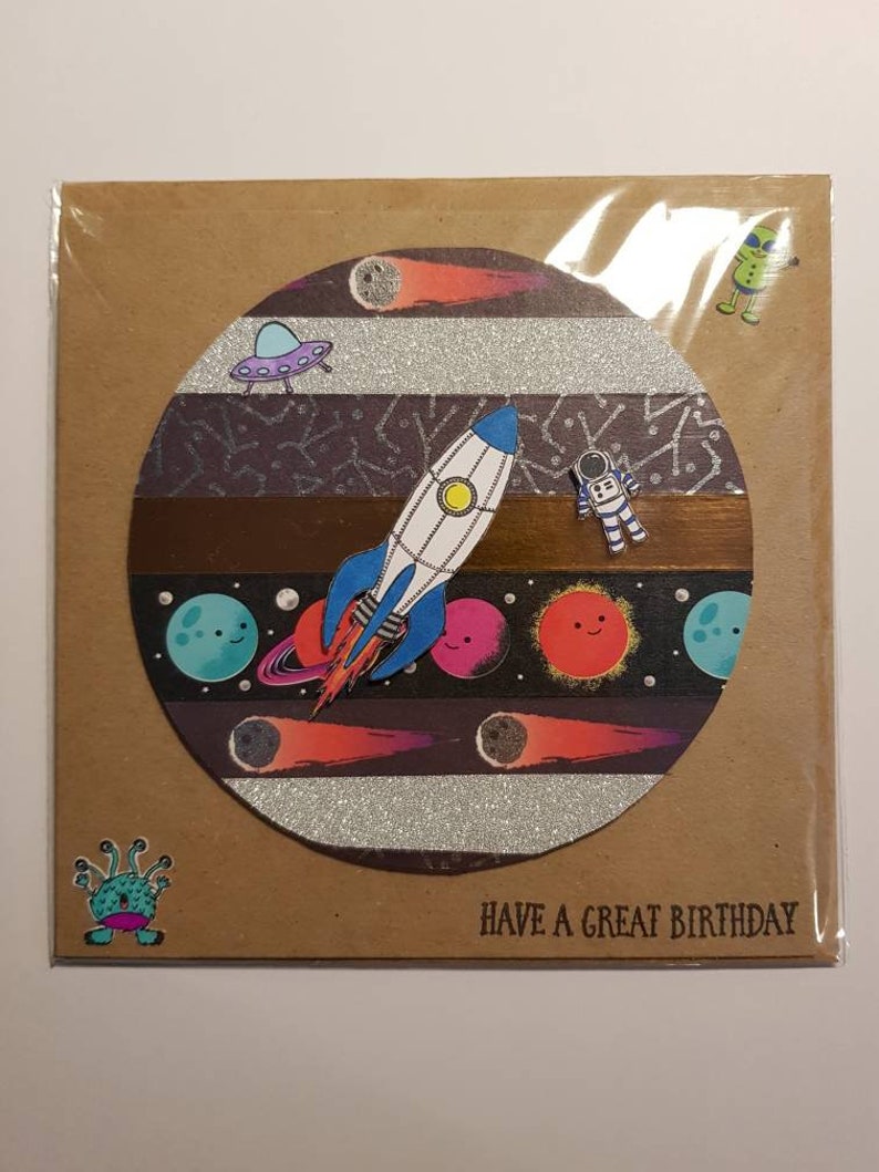 Op de afbeelding: Een bruine kaart met een cirkel in het midden. De cirkel is gevuld met een ruimte sc&egrave;ne met een raket, planeten, sterren en een UFO. De tekst "HAVE A GREAT BIRTHDAY" staat onderaan de kaart.