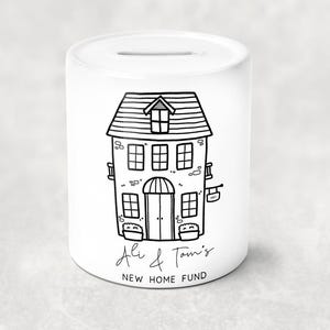 May include: White ceramic money bank featuring a black line drawing of a house. The house illustration includes windows, a door, and a sign that says "ART". The text "Ali & Tom's NEW HOME FUND" is written below the house.