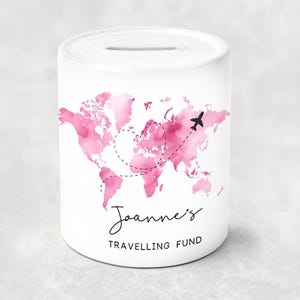 May include: White ceramic money box with a slot on top. It features a pink watercolour world map design with a dotted airplane flight path. The text "Joanne's Travelling Fund" is written in black script.