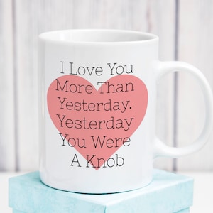 May include: White ceramic mug with a handle, featuring the text "I Love You More Than Yesterday. Yesterday You Were A Knob" in black, with a pink heart graphic. The mug is sitting on a light blue box.