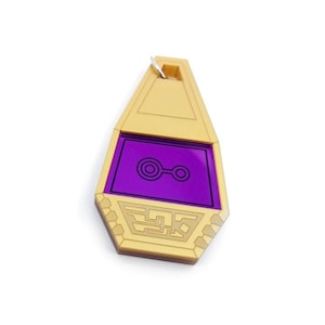 May include: A gold-colored Digimon-style keychain with a purple screen displaying a symbol. The keychain is shaped like a teardrop and has a silver loop at the top.