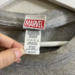 Puede incluir: Camiseta gris con una etiqueta de logo de Marvel. La etiqueta tiene el texto "MARVEL" en letras rojas y blancas. La etiqueta tambi&eacute;n tiene el texto "marvelsco" en letras negras. La etiqueta tiene instrucciones de cuidado e informaci&oacute;n de tama&ntilde;o. La talla es mediana.