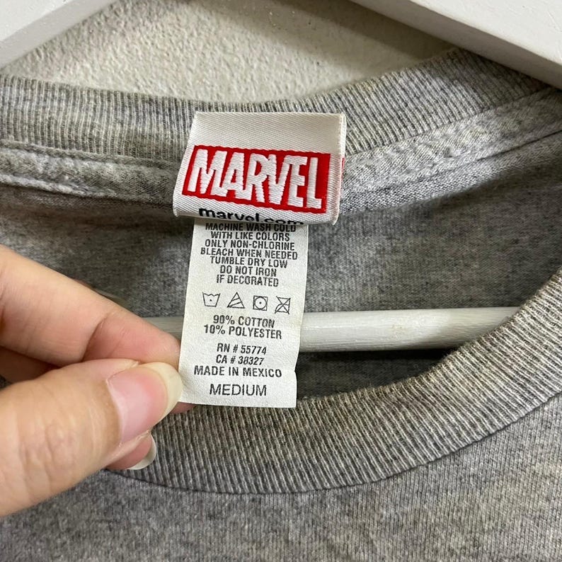 Puede incluir: Camiseta gris con una etiqueta de logo de Marvel. La etiqueta tiene el texto "MARVEL" en letras rojas y blancas. La etiqueta tambi&eacute;n tiene el texto "marvelsco" en letras negras. La etiqueta tiene instrucciones de cuidado e informaci&oacute;n de tama&ntilde;o. La talla es mediana.