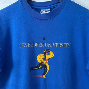 May include: Royal blue t-shirt with the text "DEVELOPER UNIVERSITY" in gold lettering and a black apple logo. The shirt features a colorful graphic of a running figure with a yellow body and black and red details.