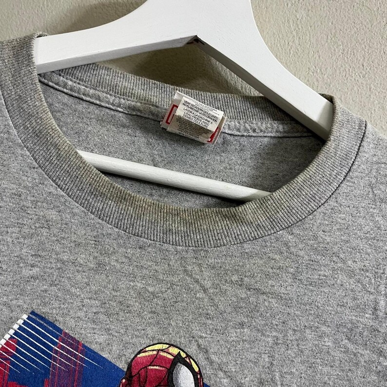 Puede incluir: Camiseta gris con un gr&aacute;fico de Spider-Man en un traje rojo y azul. La camiseta tiene una etiqueta que dice "100% algod&oacute;n, Hecho en EE. UU., Lavar a m&aacute;quina con agua fr&iacute;a, Secar en secadora a baja temperatura, No usar blanqueador".