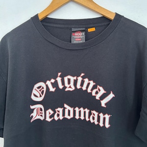 May include: Black t-shirt with the text "original Deadman" in a white and red gothic font. The shirt is hanging on a wooden hanger. The brand label "SHOOT" is visible at the neckline.