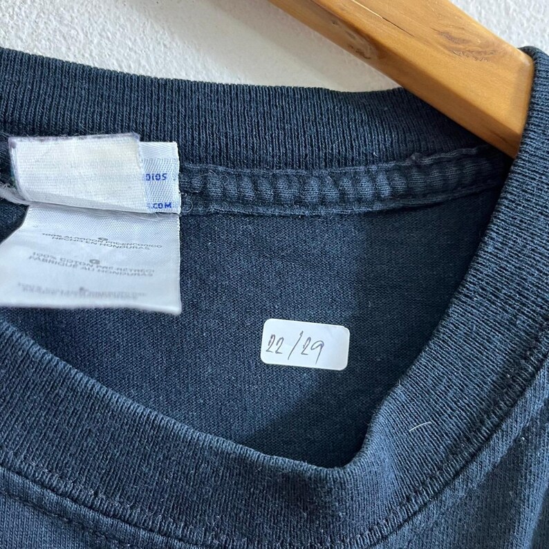 Op de afbeelding: Zwart T-shirt met een wit label met de tekst "studios.com" en een wit label met het nummer "22/29".