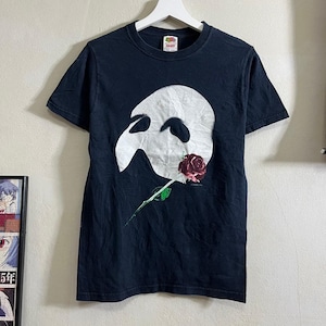 以下が含まれることがあります： 黒いTシャツに、オペラ座の怪人のマスクと赤いバラの白いグラフィックがプリントされています