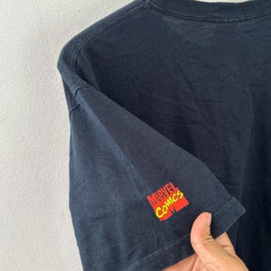 May include: Black t-shirt with the Marvel Comics logo in red and yellow on the sleeve. The shirt is made of a soft material and has a crew neck.