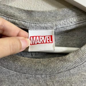 Puede incluir: Una camiseta gris con una etiqueta blanca que dice "MARVEL" en letras rojas.