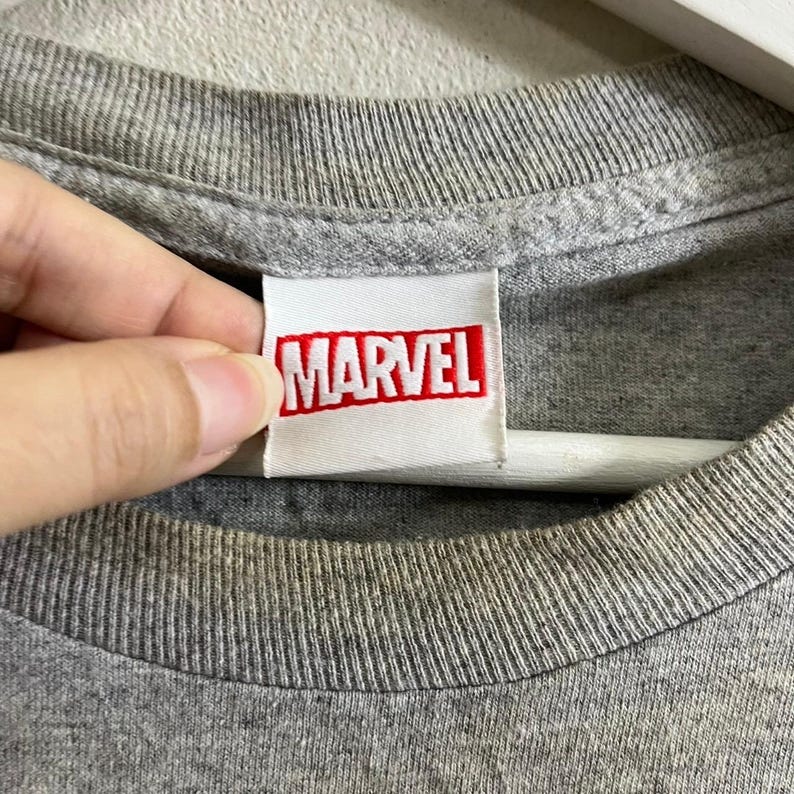Puede incluir: Una camiseta gris con una etiqueta blanca que dice "MARVEL" en letras rojas.