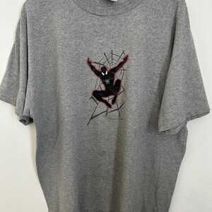 Puede incluir: Una camiseta gris con un gr&aacute;fico de Spider-Man negro, detalles en rojo y un dise&ntilde;o de telara&ntilde;a. El superh&eacute;roe se muestra en una pose din&aacute;mica con los brazos extendidos. La camiseta est&aacute; hecha de un material suave y tiene un cuello redondo cl&aacute;sico.