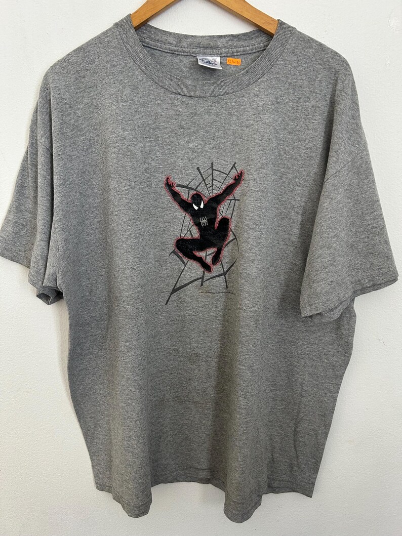 Puede incluir: Una camiseta gris con un gr&aacute;fico de Spider-Man negro, detalles en rojo y un dise&ntilde;o de telara&ntilde;a. El superh&eacute;roe se muestra en una pose din&aacute;mica con los brazos extendidos. La camiseta est&aacute; hecha de un material suave y tiene un cuello redondo cl&aacute;sico.
