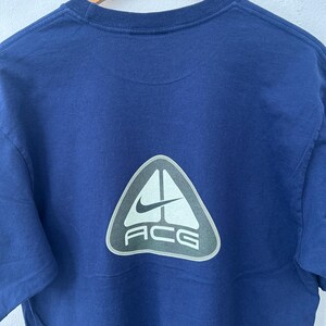 May include: Navy blue t-shirt with a white and gray triangle logo on the back. The logo features the Nike swoosh and the letters "ACG". The shirt is hanging on a wooden hanger.