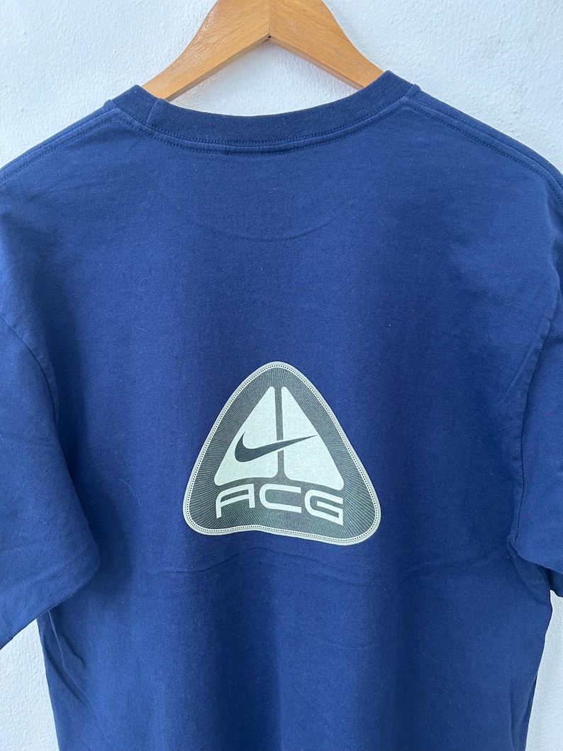 May include: Navy blue t-shirt with a white and gray triangle logo on the back. The logo features the Nike swoosh and the letters "ACG". The shirt is hanging on a wooden hanger.